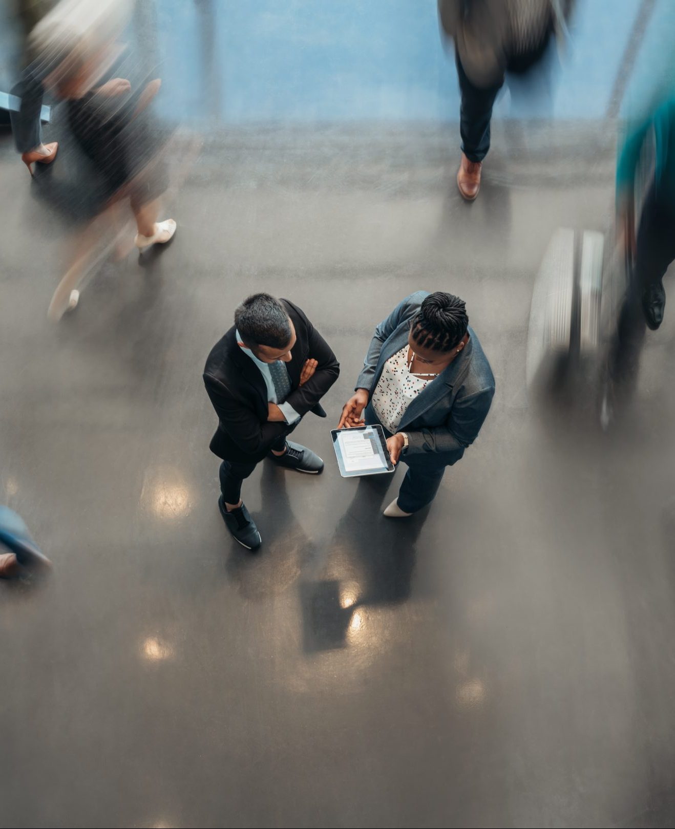 Two business people standing in the lobby of an office looking at a tablet while people are walking past in a blur